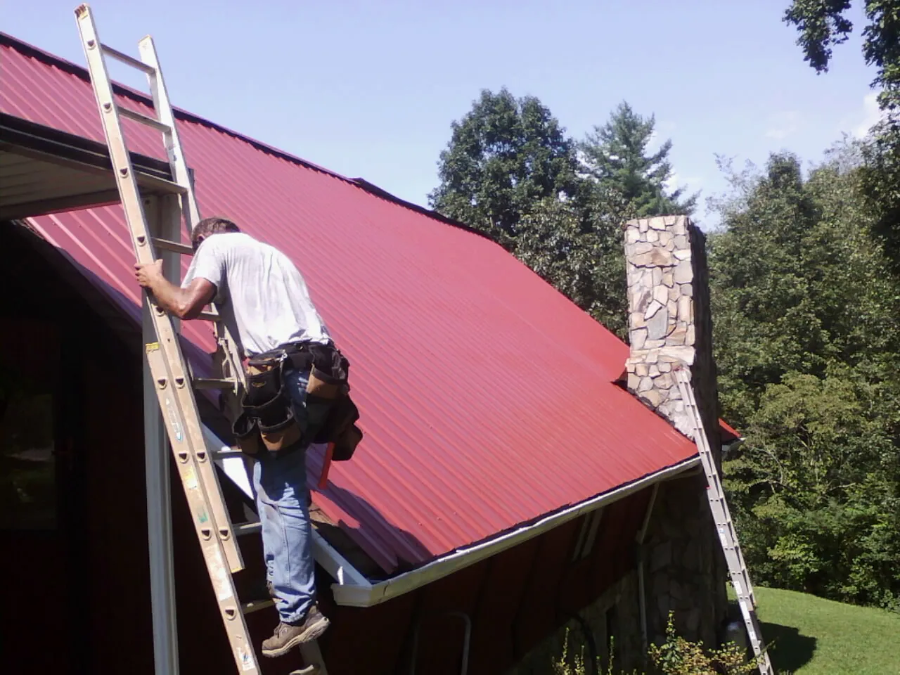 Roofer on a ladder beside a red metal roof and stone chimney — Soffit & Fascia Repair by Roofing Brothers in Bee